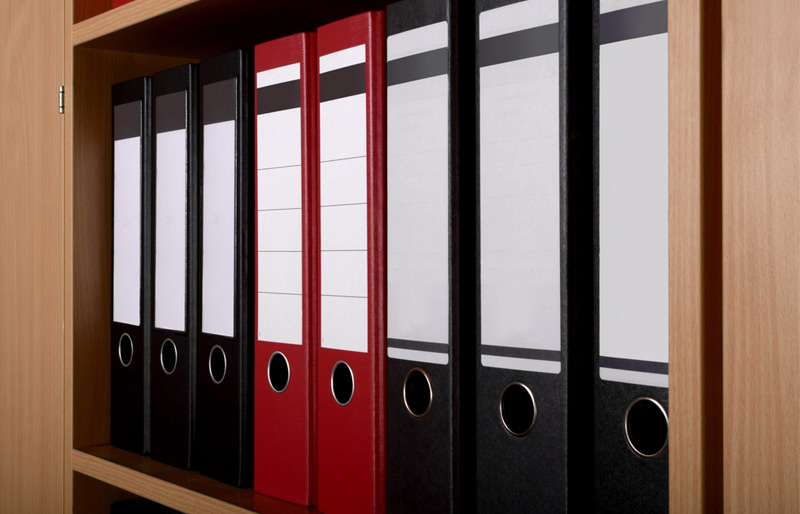 A row of eight loose-leaf binders sits on a shelf. All are black except for the two in the middle, which are red.
