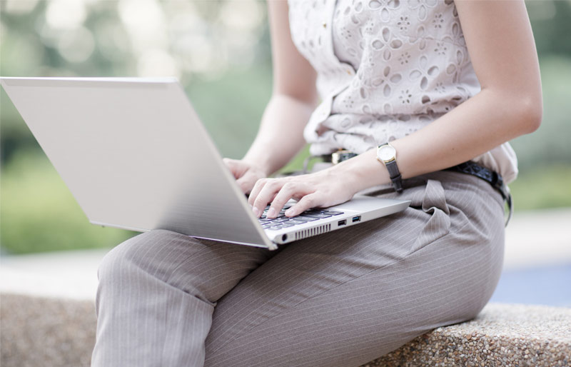 Assise sur un muret en béton dans un parc, une femme travaille sur un ordinateur posé sur ses genoux.