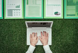 Close up of person's hands using a laptop computer on grassy surface beside pages with reports.