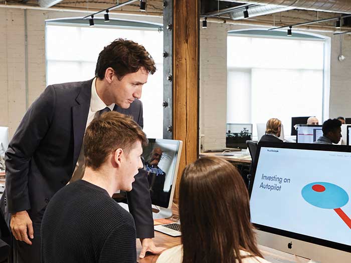 Le PM Justin Trudeau, regardant par-dessus l'épaule d'un employé de Wealthsimple, devant un écran d'ordinateur