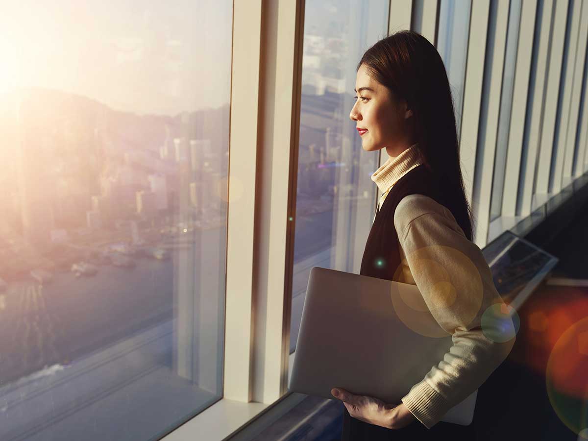 Young woman holding laptop computer, while standing in office interior and looking out of big window with city view