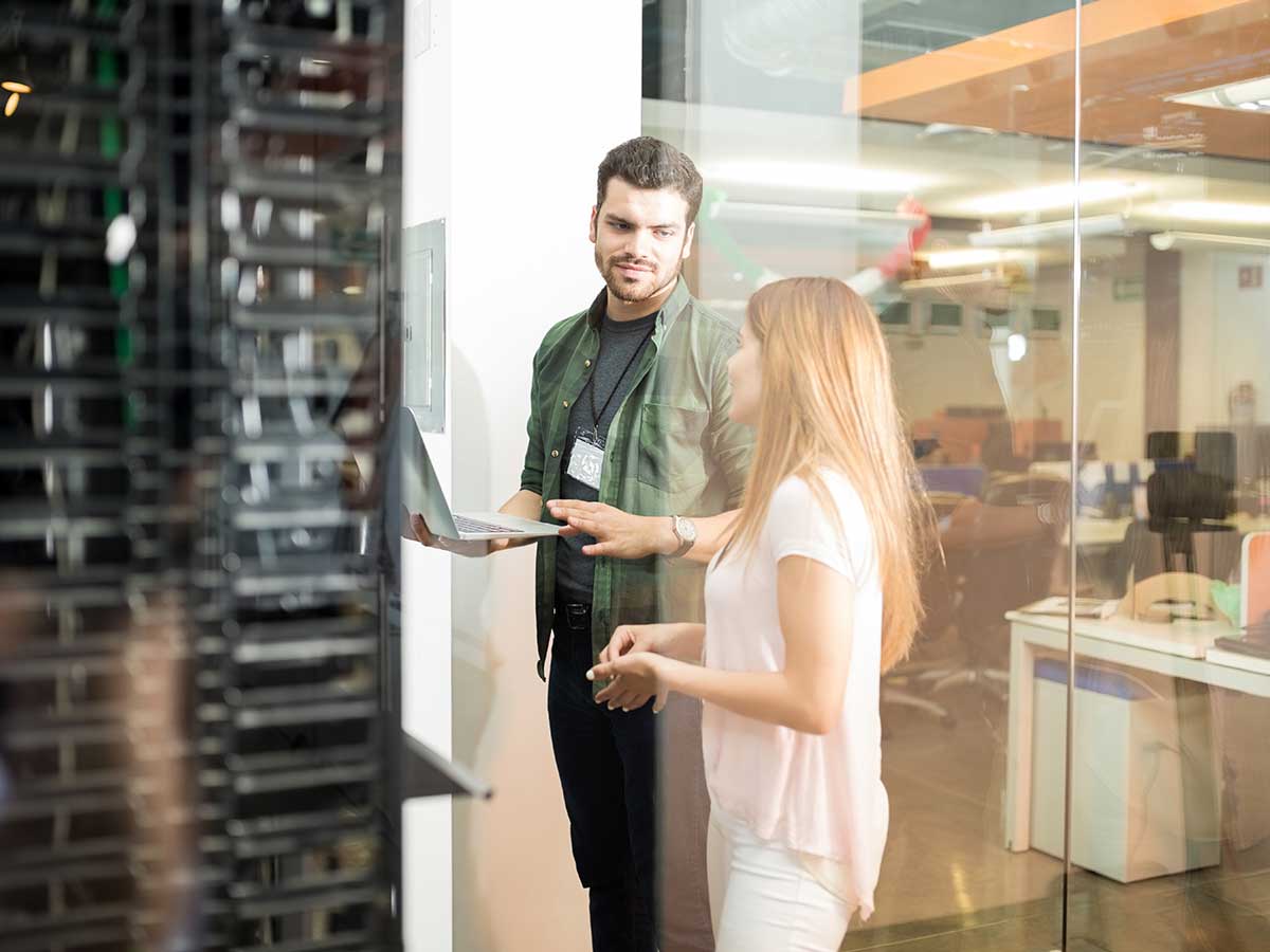 Two employees standing in server room using a laptop, with the office in background