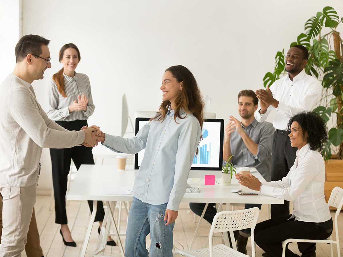Boss shaking hand of young woman, while business team applauding in background