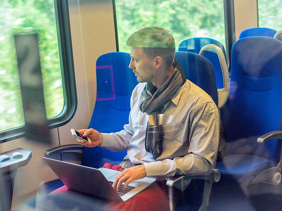 Young man on train, using smart phone and a laptop, while commuting