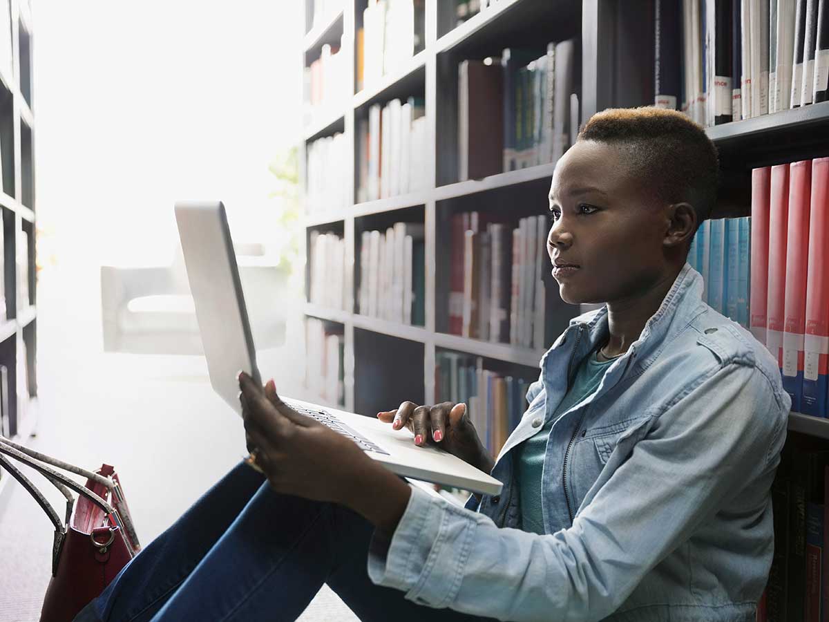 Young lady using laptop between library bookshelves to study