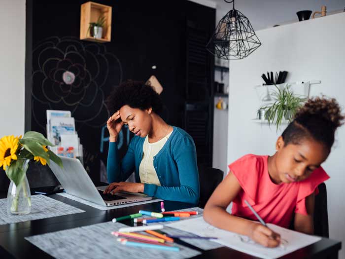 Young mother working on laptop at table while daughter is sharing space being home schooled