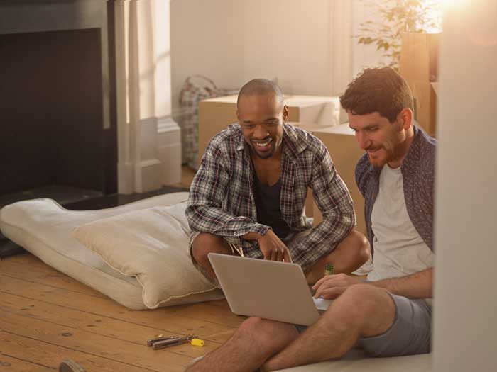 Two men sitting on living room floor of new home looking at laptop with packed boxes behind them