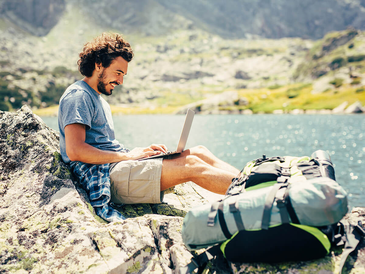 Randonneur masculin se reposant et utilisant un ordinateur portable sur un rocher près d'un lac dans la montagne.