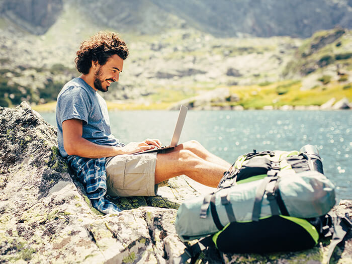 Male hiker resting and using a laptop on a rock near a lake in the mountain.
