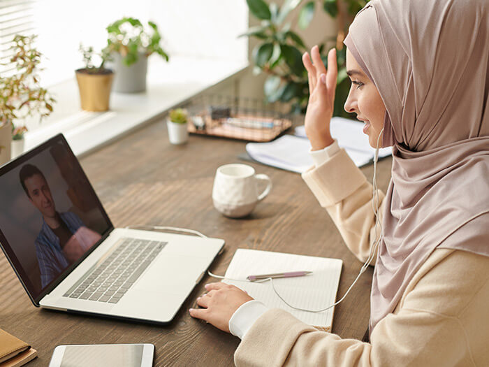 Muslim woman wearing headwrap sitting at desk having online meeting greeting her colleague