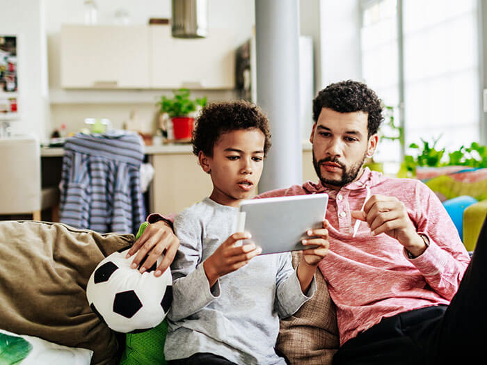 A father and son are sitting together on a couch, looking at a tablet