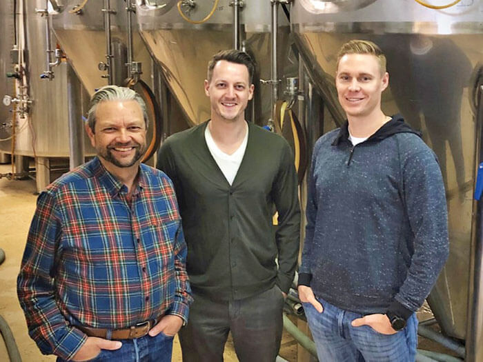 Three men stand in front of large silver tanks of beer