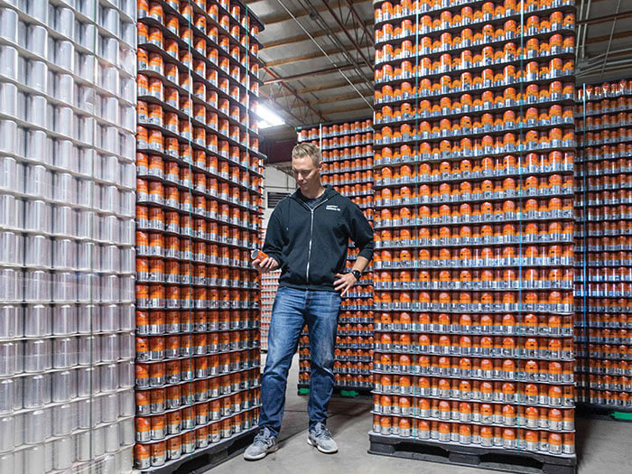 A man holds a can of beer while standing in front of stacked skids of beer cans