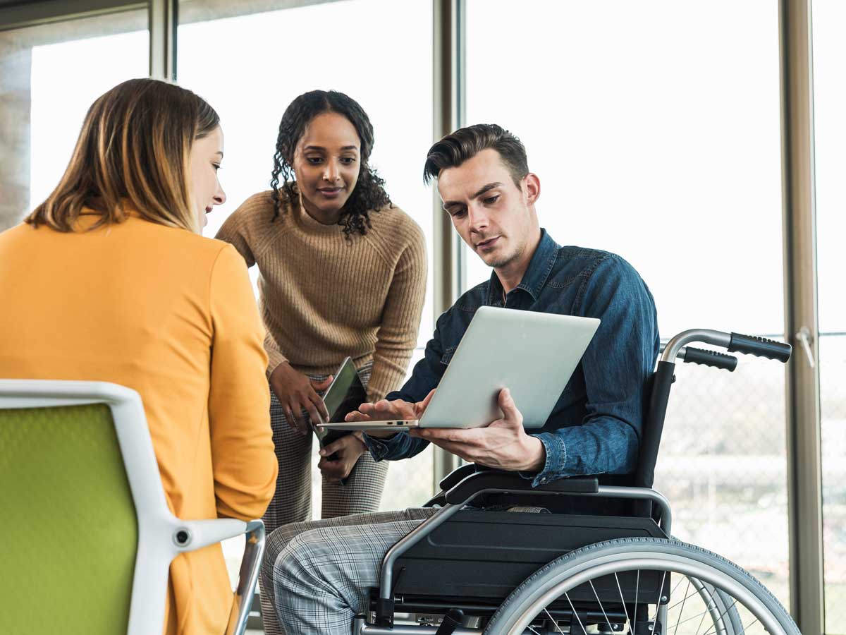 Young businessman in wheelchair showing laptop to colleague in office 