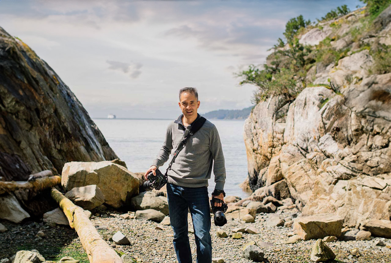 Ron Yue se tient sur des rochers au bord de l’eau, son matériel de photographie à la main.