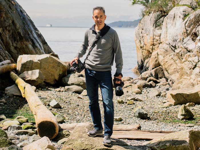 Ron Yue stands on a rocky shore with with photography equipment in hand