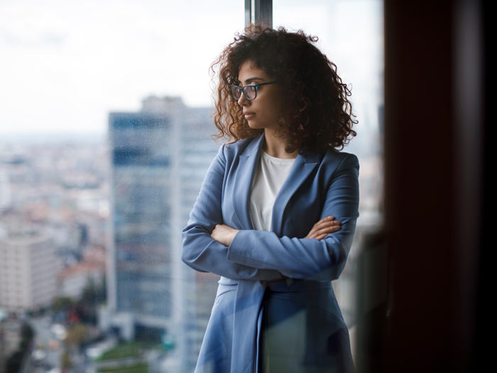Young thoughtful businesswoman wearing a suit standing in her office and looking through window 