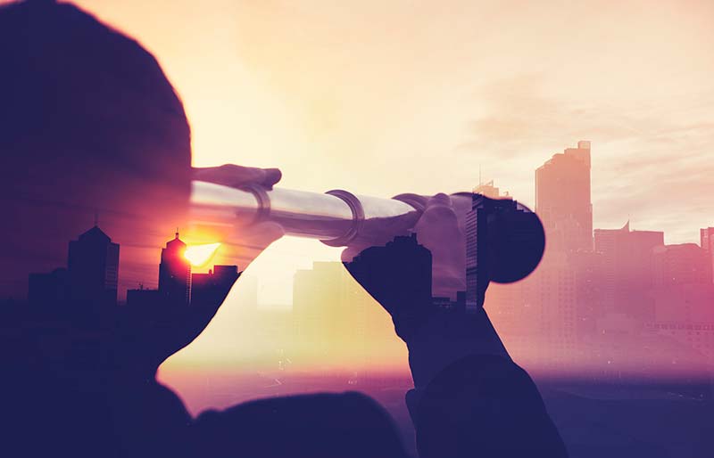 A silhouette of a business man looking at the city through a telescope during sunset.