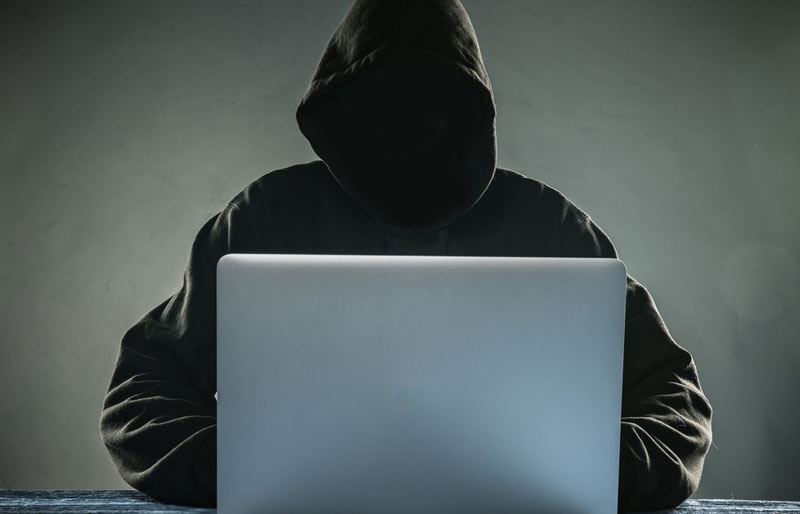 A close-up of a person in a hooded sweat shirt on a laptop, face in the shadows.
