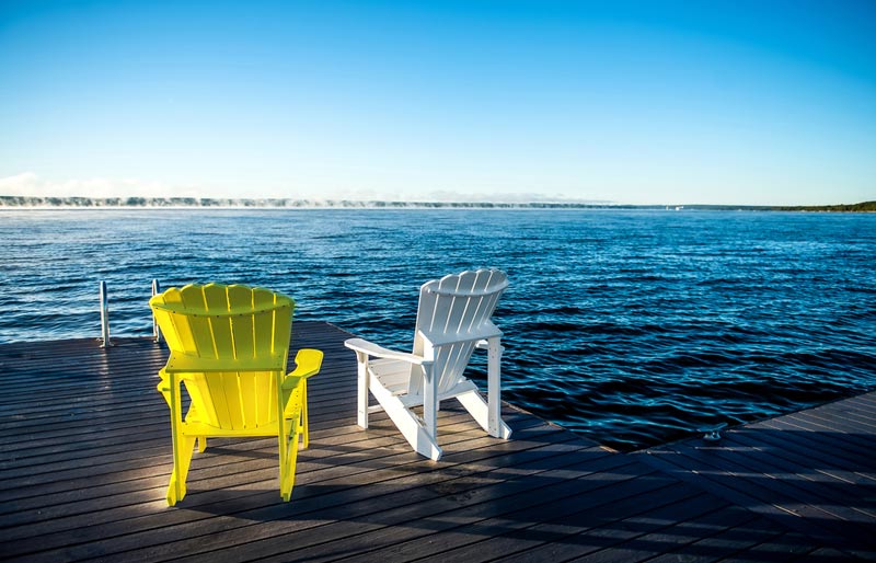 A photograph of two Muskoka chairs sitting on a deck looking out over a beautiful lake.