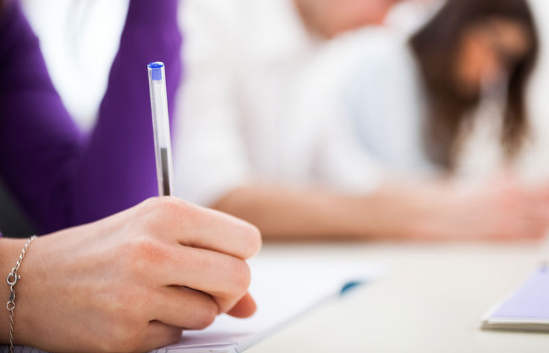 Gros plan sur la main d’une femme installée à une table en train d’écrire au stylo à bille sur une feuille blanche. À l’arrière-plan, d’autres personnes sont également en train d’écrire.