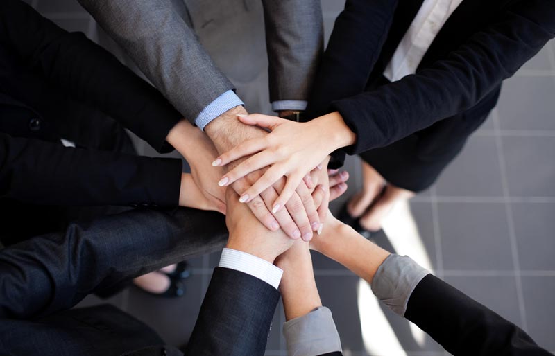 An aerial photo shows businesspeople standing in a circle with their arms reaching in towards the middle of the circle and hands stacked on top of each other.