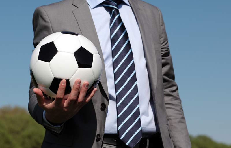A man wearing a suit stands outdoors in front of a grassy hill set against a blue sky and holds a soccer ball in his right hand.