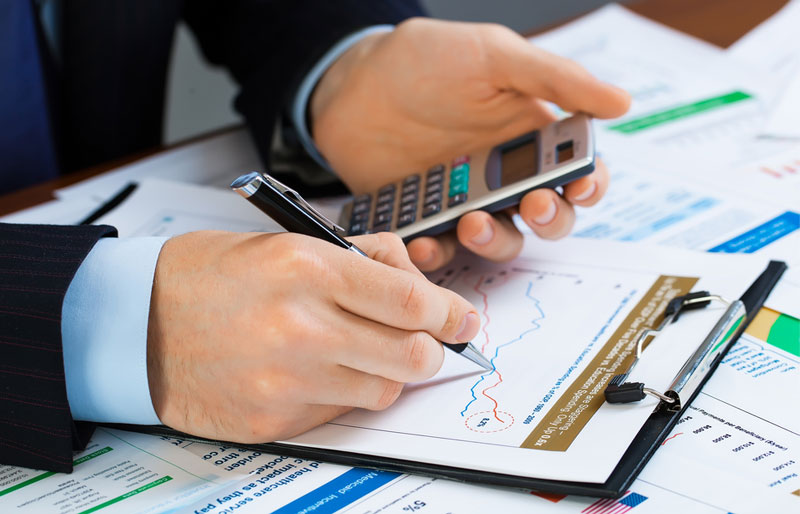 A businessman holds a calculator in one hand and traces a graph with his pen on the printout in front of him.