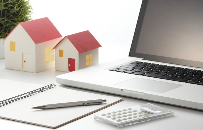 Deux maisons miniatures, un ordinateur portable, une calculatrice, et un bloc-notes sur lequel repose un stylo.
