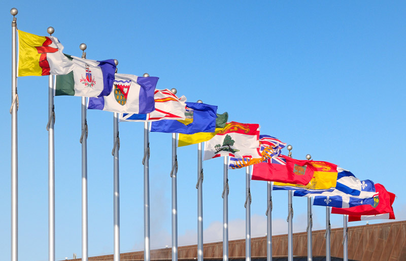 Flags of the Canadian provinces and territories are seen flying in the wind atop a series of flagpoles under a blue sky.