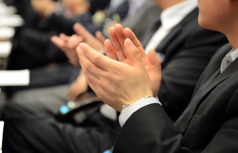 A close-up of a businessman’s hands clapping. He is part of a larger audience of businesspeople.
