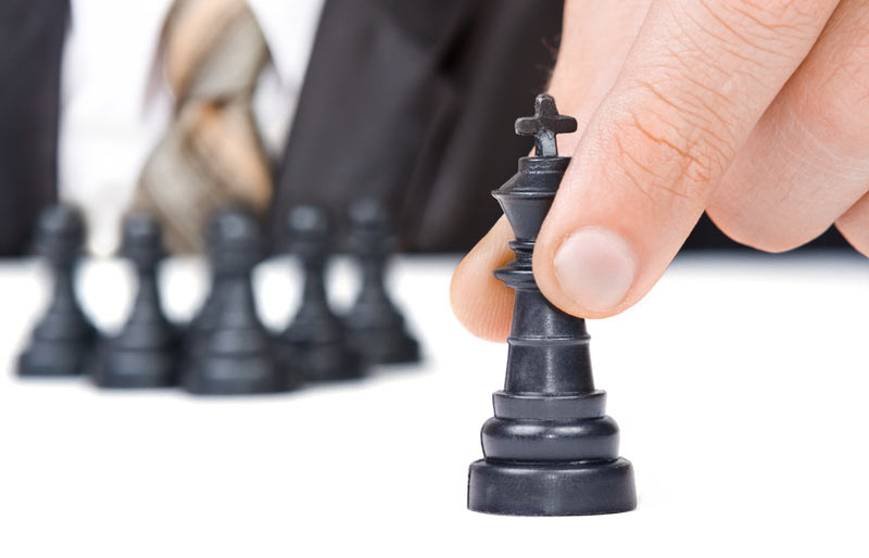 A businessman holds the figure of a black king from a chess set while six pawns are gathered in the background.