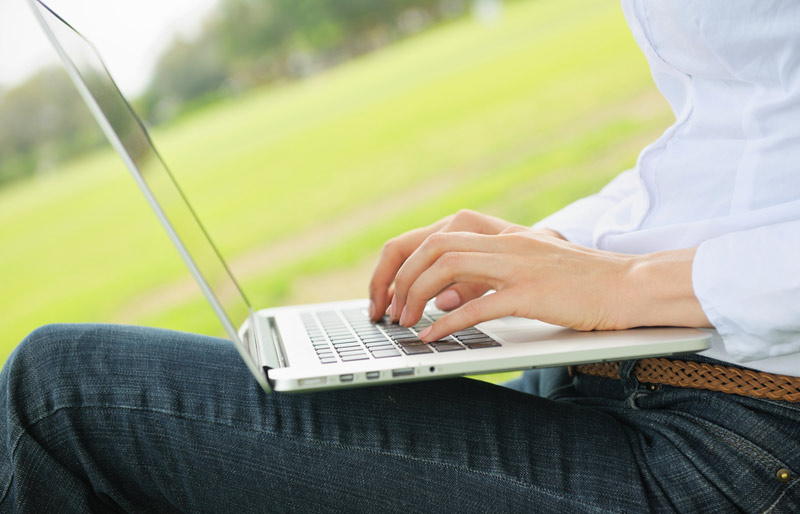 A woman wearing jeans and a white shirt types on a laptop outdoors in a park setting.