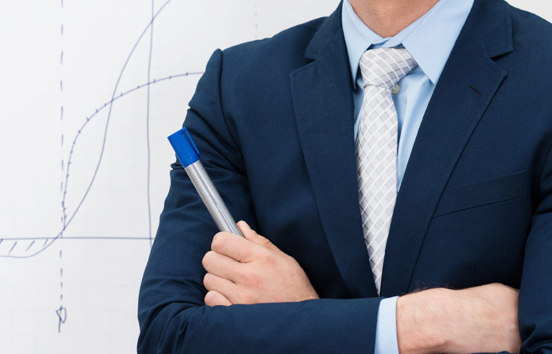 A businessman with arms crossed holds a marker. He is standing in front of a whiteboard on which is drawn a graph. 