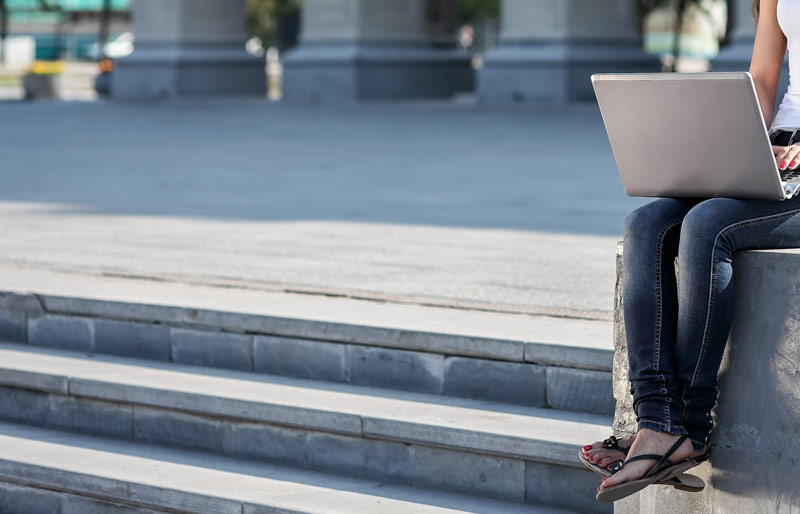 A young woman wearing jeans has a laptop on her lap and is perched on a concrete wall next to some stone steps in an outdoor setting. 