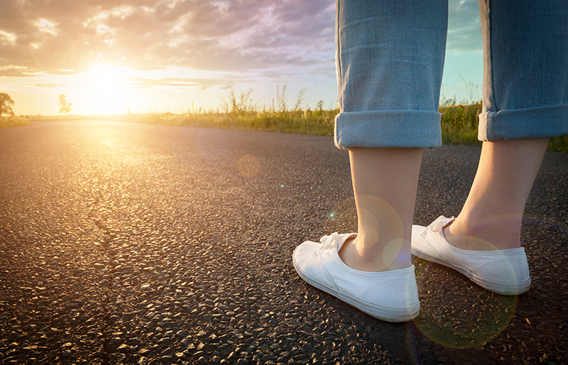 feet of a person standing on a road, facing the sun setting at end of the road