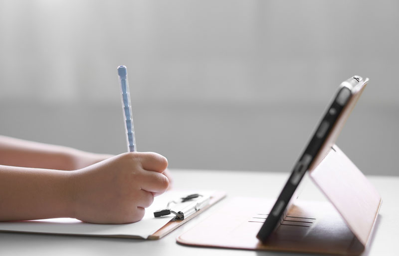 Gros plan sur les mains d’un enfant, qui écrivent sur un carnet de notes, devant une tablette numérique.