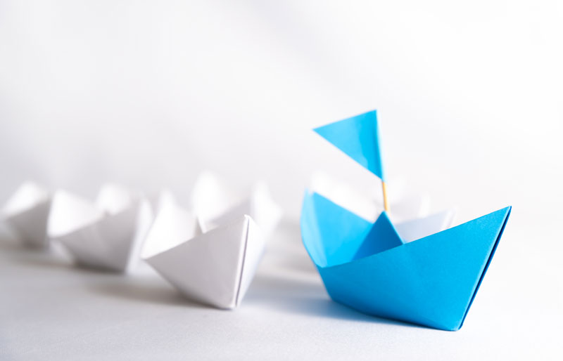 Blue paper boat leading a row of white paper boats.