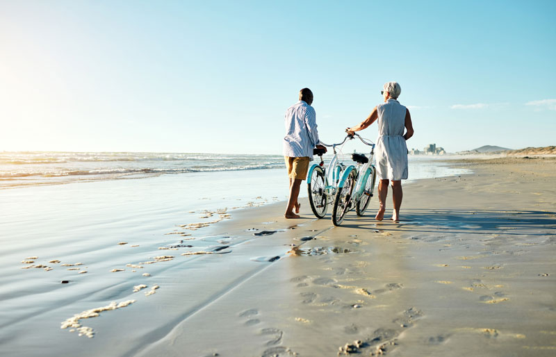 Two people walk on a sandy beach with a bicycle.
