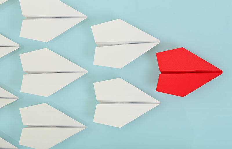 A red paper plane leading a group of white paper planes. 