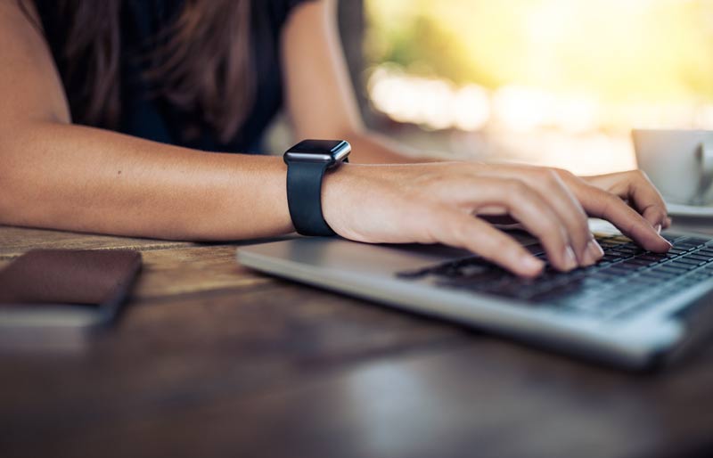 Gros plan sur les mains d’une femme portant une montre intelligente qui travaille sur un ordinateur portable.