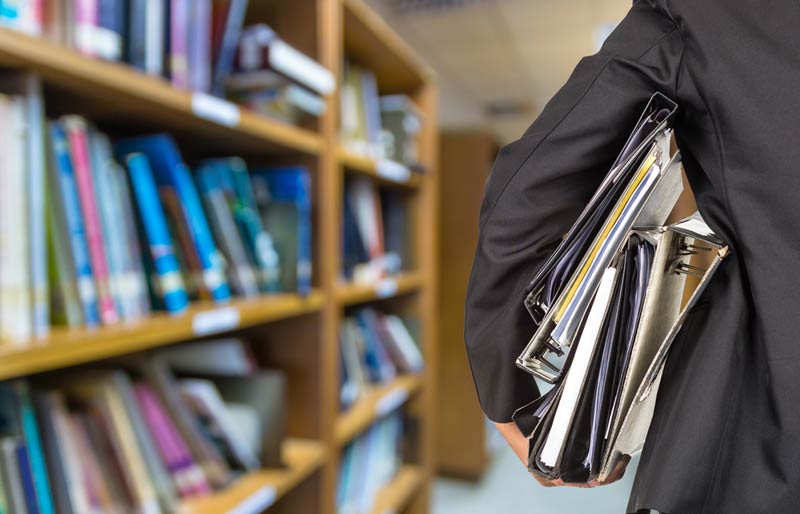 A close-up of a business professional holding a stack of binders and standing in a library. 