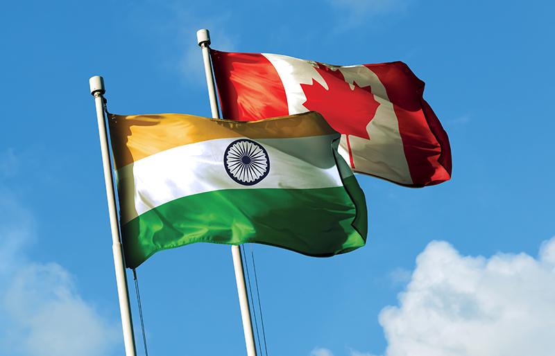 Flags for both India and Canada waving together in the breeze against a blue sky