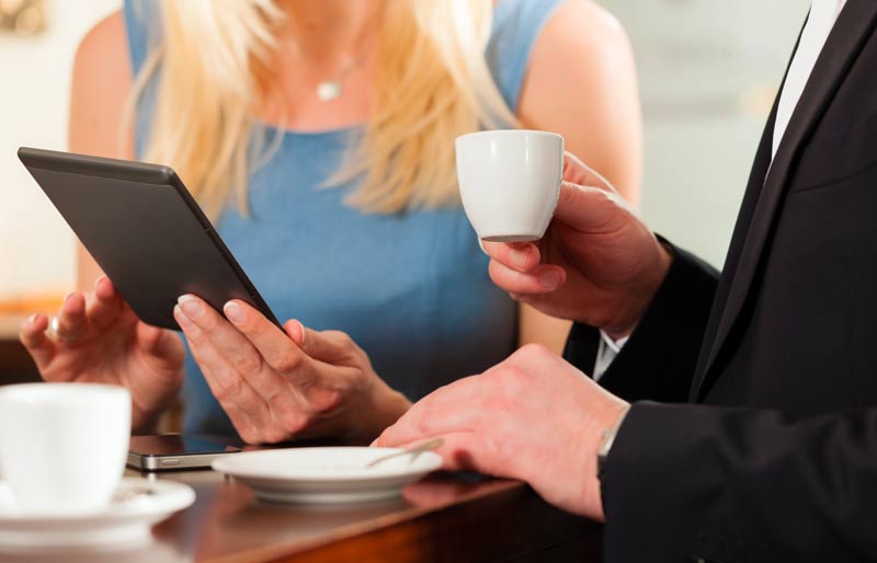 Deux professionnels assis à une table, dont une femme qui consulte une tablette et un homme qui tient une tasse à café.