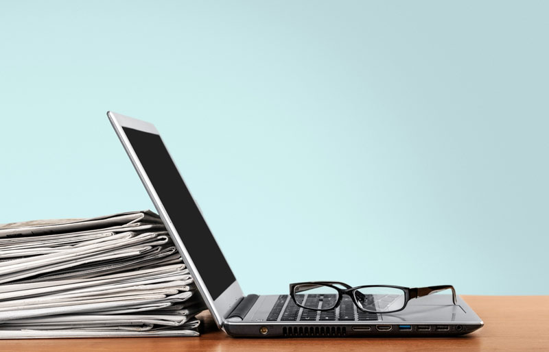 Un ordinateur portable, une pile de journaux et une paire de lunettes sont posés sur un bureau.