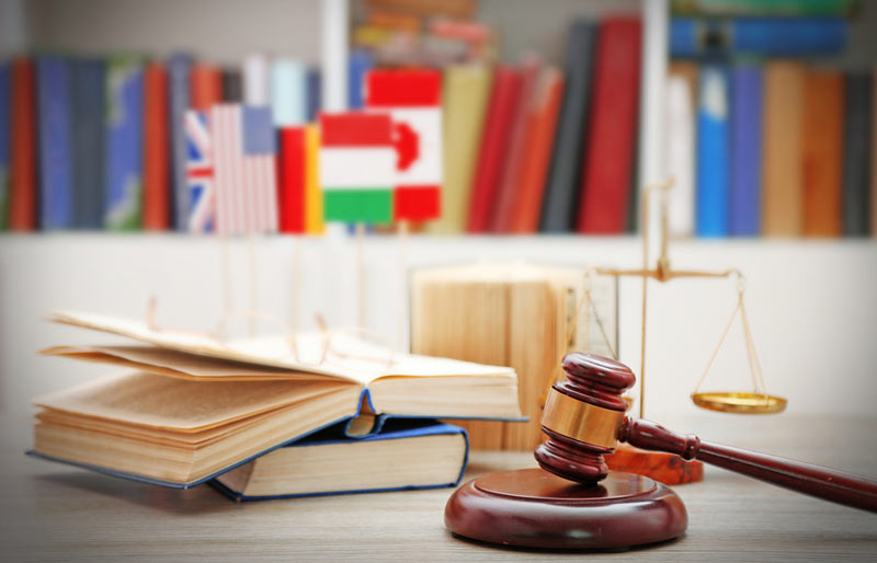 A close-up photograph of a court justice gavel on a desk with law books and in the background various countries flags including Canada.