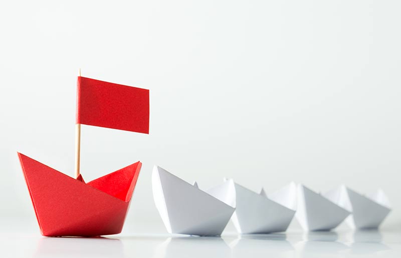 Petit bateau en papier rouge suivi de quatre plus petits bateaux en papier blanc. 