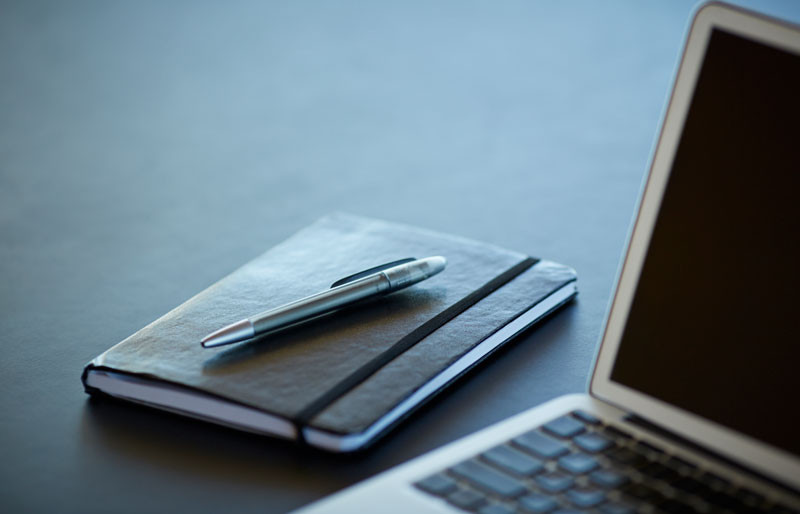 Cadrage sur un calepin de notes, un stylo et un ordinateur portable posés sur un bureau de travail