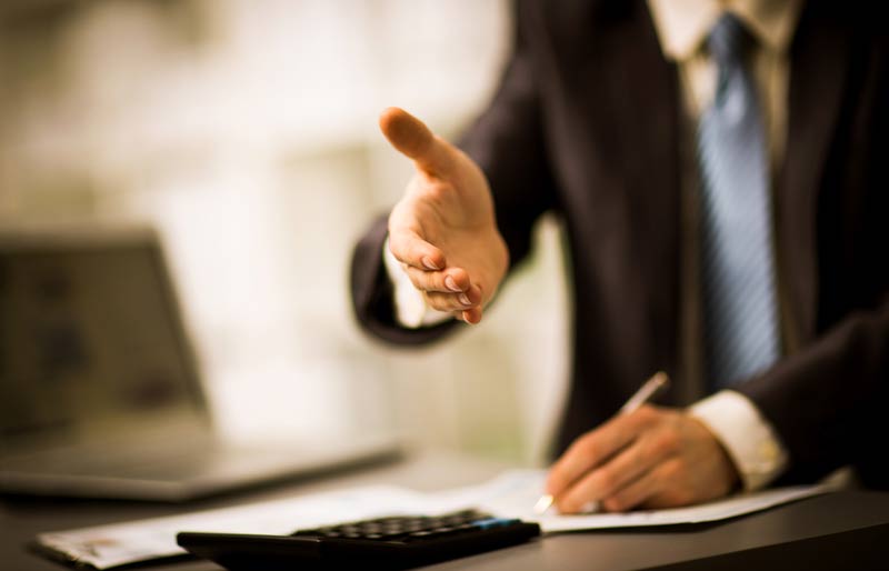 Un professionnel assis à un bureau signe un contrat de la main gauche et tend la main droite à quelqu’un d’autre pour lui serrer la main. 