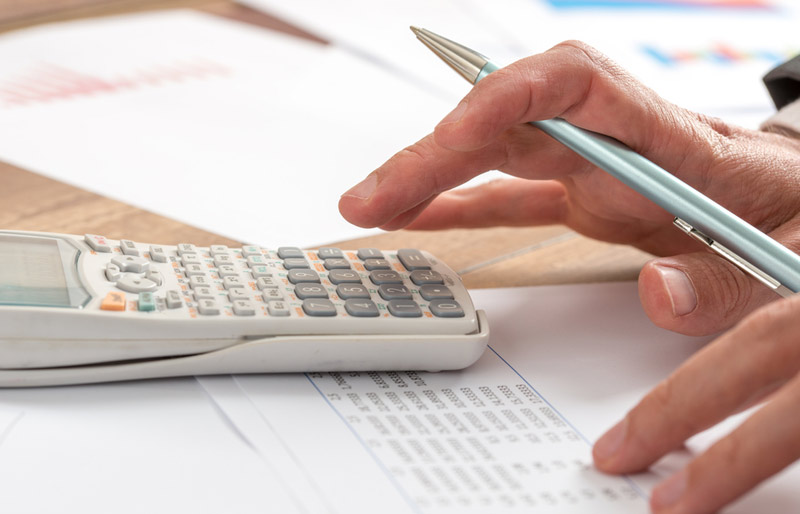 A close-up of a business professional holding a pen and working on a calculator and spread sheets.